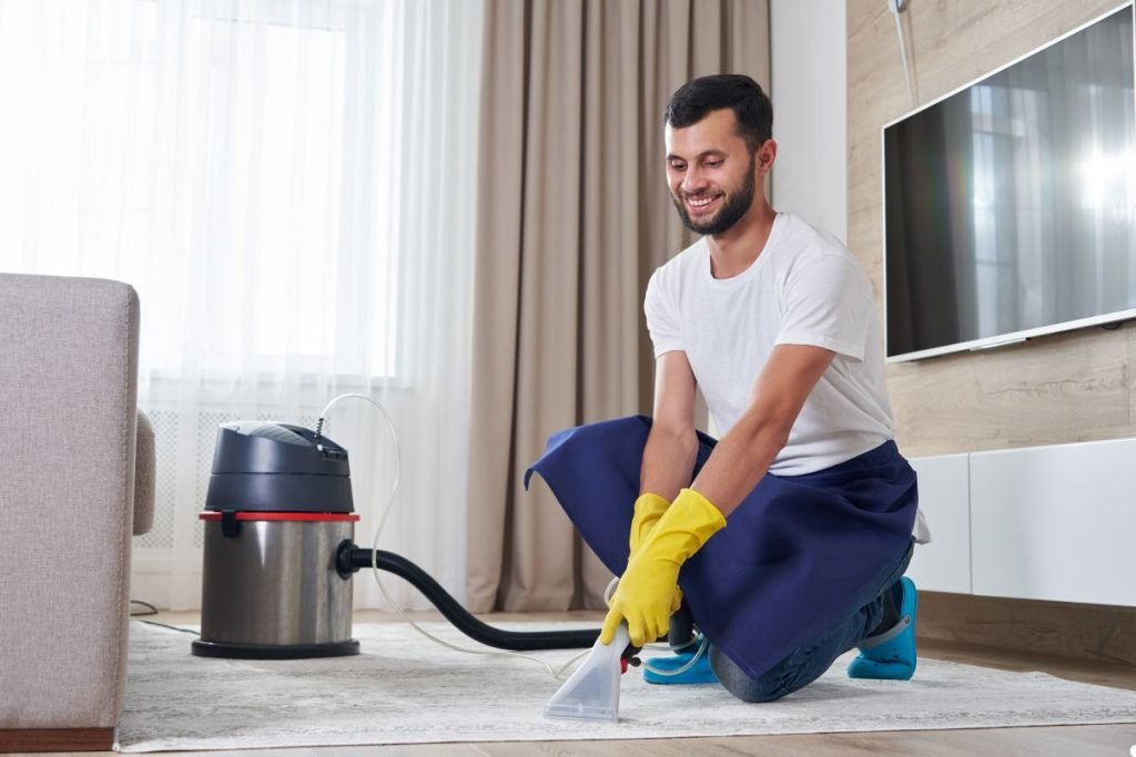 A man wearing a blue shirt and yellow gloves is cleaning a carpet with a vacuum cleaner.