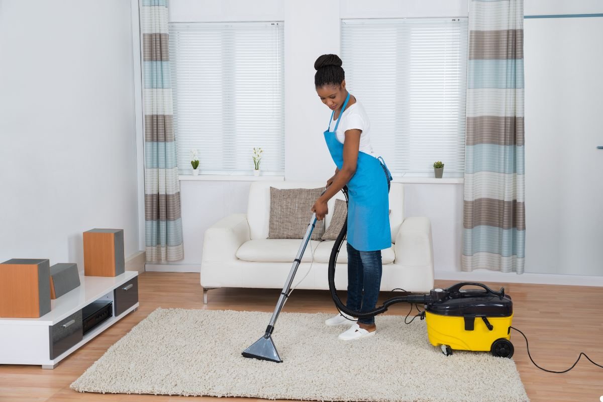 A woman wearing an apron vacuums a room, focusing on cleaning the carpeted floor.